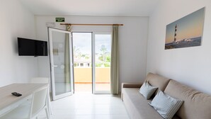 A room with a sofa, a TV, a desk, and a balcony with a view of buildings and greenery.