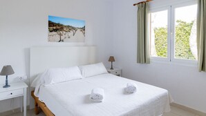 A neatly made bed with white linens and two rolled towels, a bedside table with a lamp, a nightstand, a window with curtains, and a beach scene painting on the wall.
