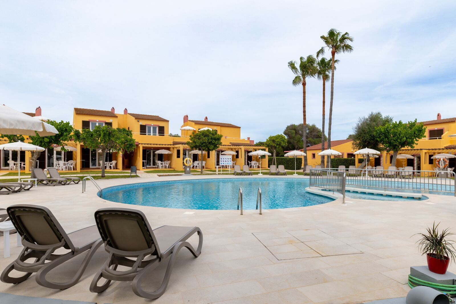 Zona de piscina amb gandules i ombrelles, envoltada d'edificis grocs amb palmeres.