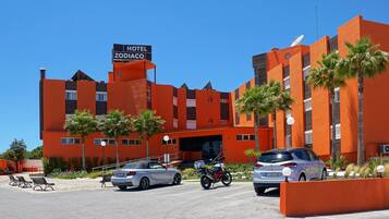 A hotel with a sign reading "HOTEL ZODIACO," featuring a modern orange facade, palm trees, and a parking area with cars and a motorcycle.