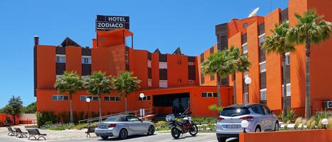 A hotel with a sign reading "HOTEL ZODIACO," featuring a modern orange facade, palm trees, and a parking area with cars and a motorcycle.