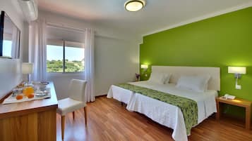 Ein Hotelzimmer mit Bett, Nachttischen, Schreibtisch mit Stuhl, Fenster mit Vorhängen und Blick auf Grünfläche.