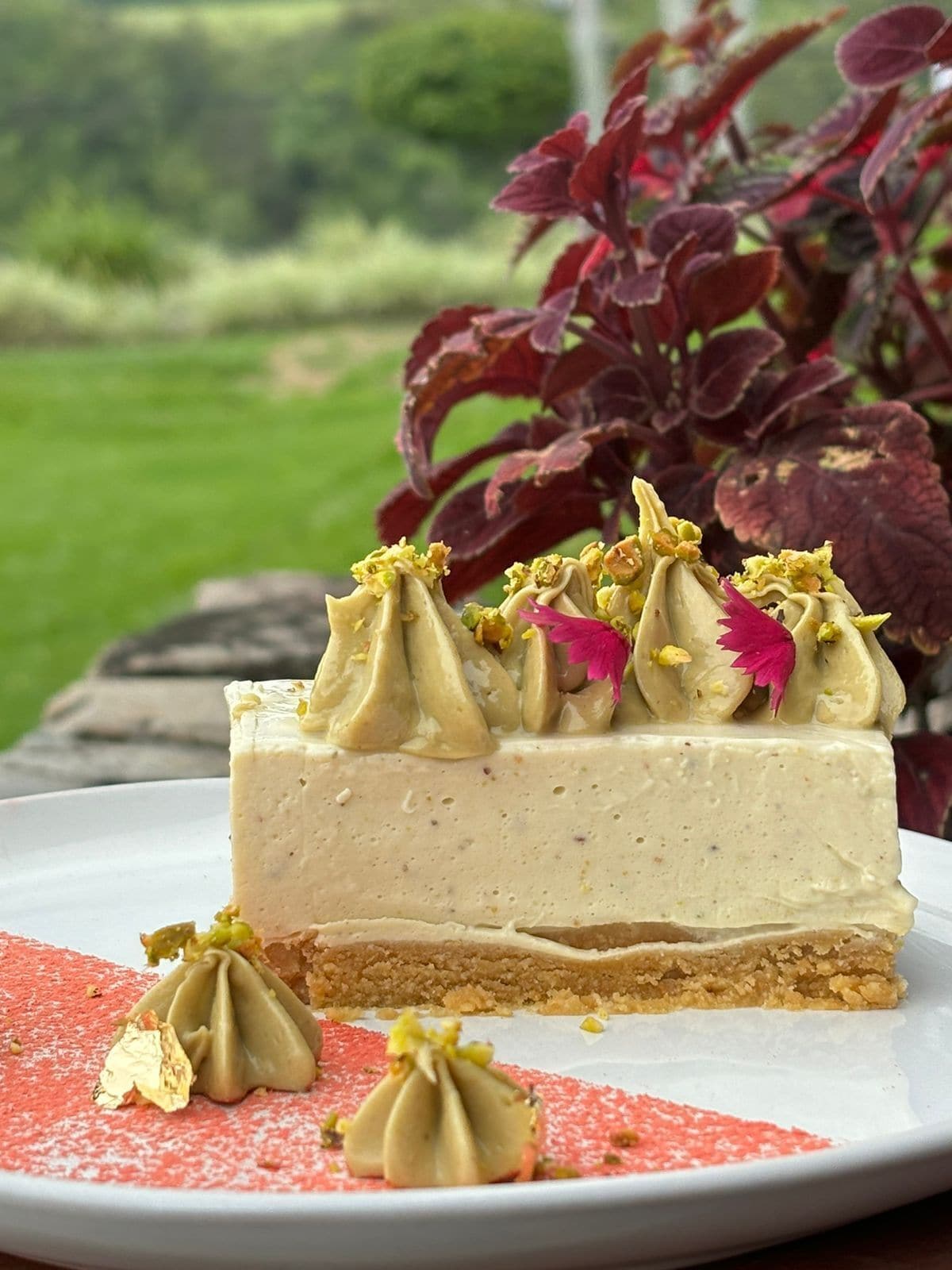 Uma fatia de cheesecake com base crocante e cobertura cremosa, decorada com flores comestíveis e pistácios, sobre um prato branco com toalha de mesa estampada em vermelho e branco.