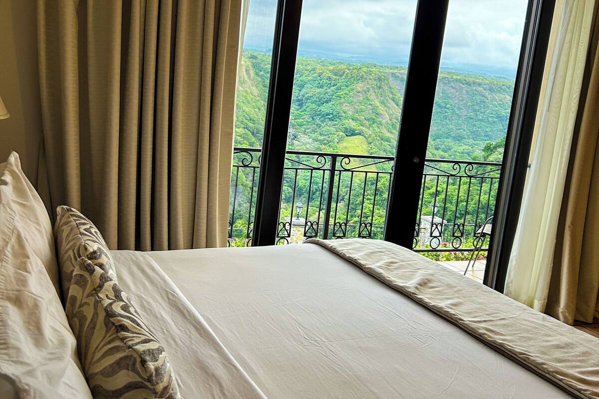 Habitación de hotel con una cama grande, cortinas color crema y vista a un paisaje verde exuberante desde un balcón.