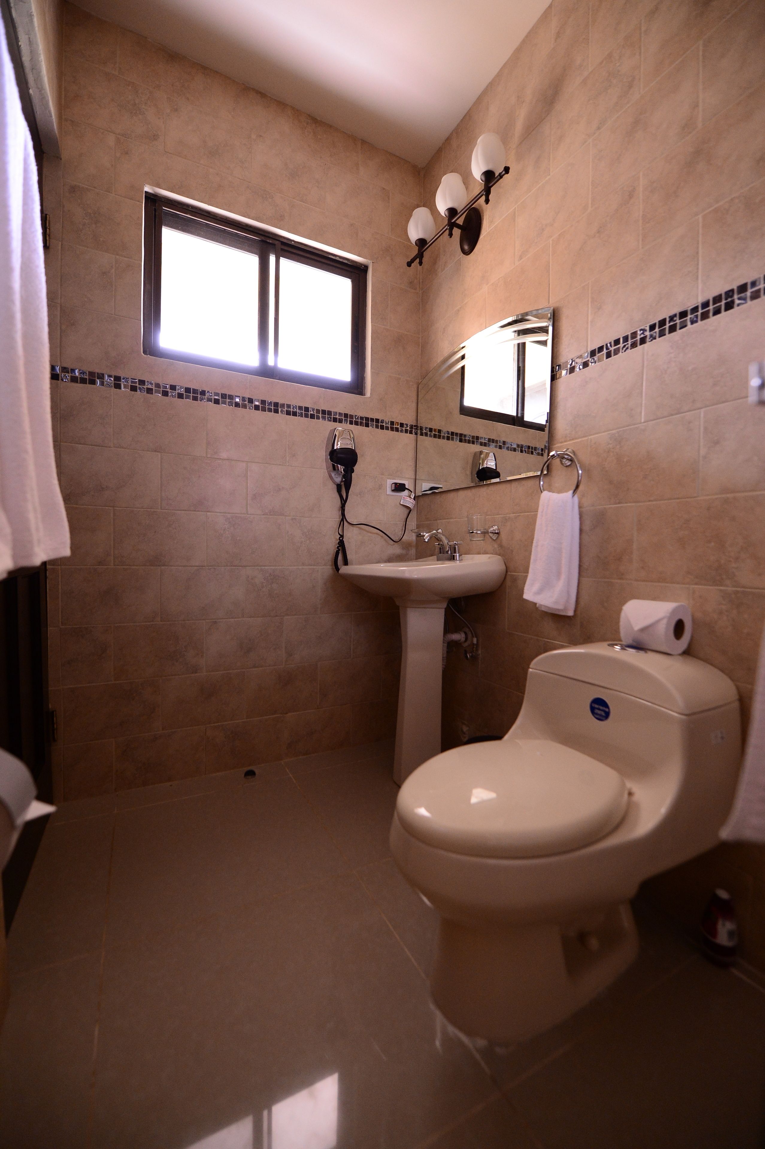 A bathroom with beige tiles, a sink, a toilet, a window, and a mirror.
