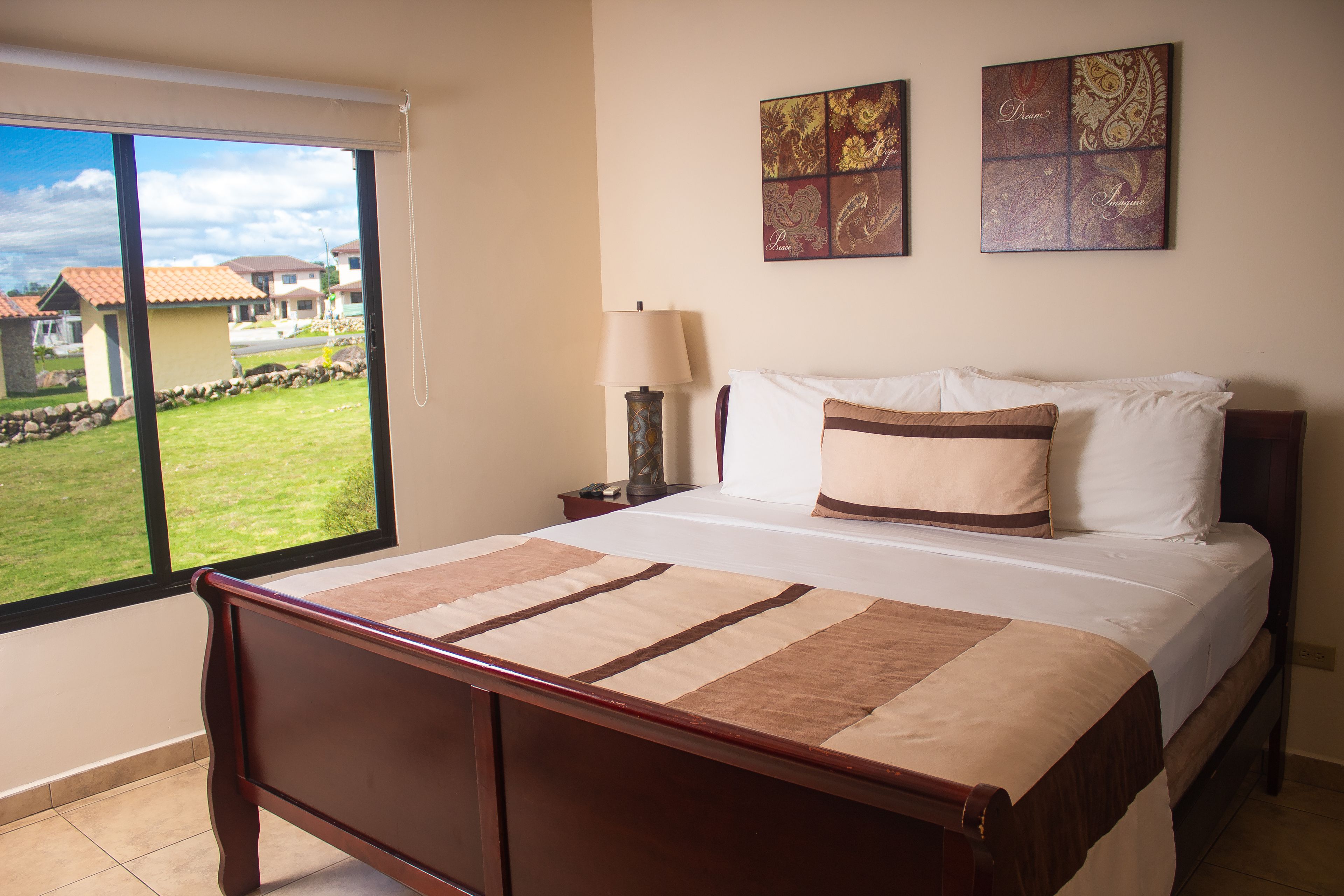 Un dormitorio con una cama grande, una mesita de noche, una lámpara y vistas a un jardín verde y casas a través de un ventanal corredera.