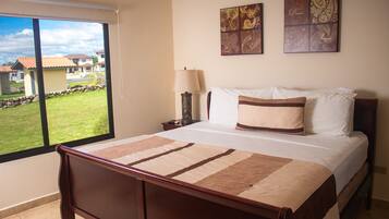 Un dormitorio con una cama grande, una mesita de noche, una lámpara y vistas a un jardín verde y casas a través de un ventanal corredera.