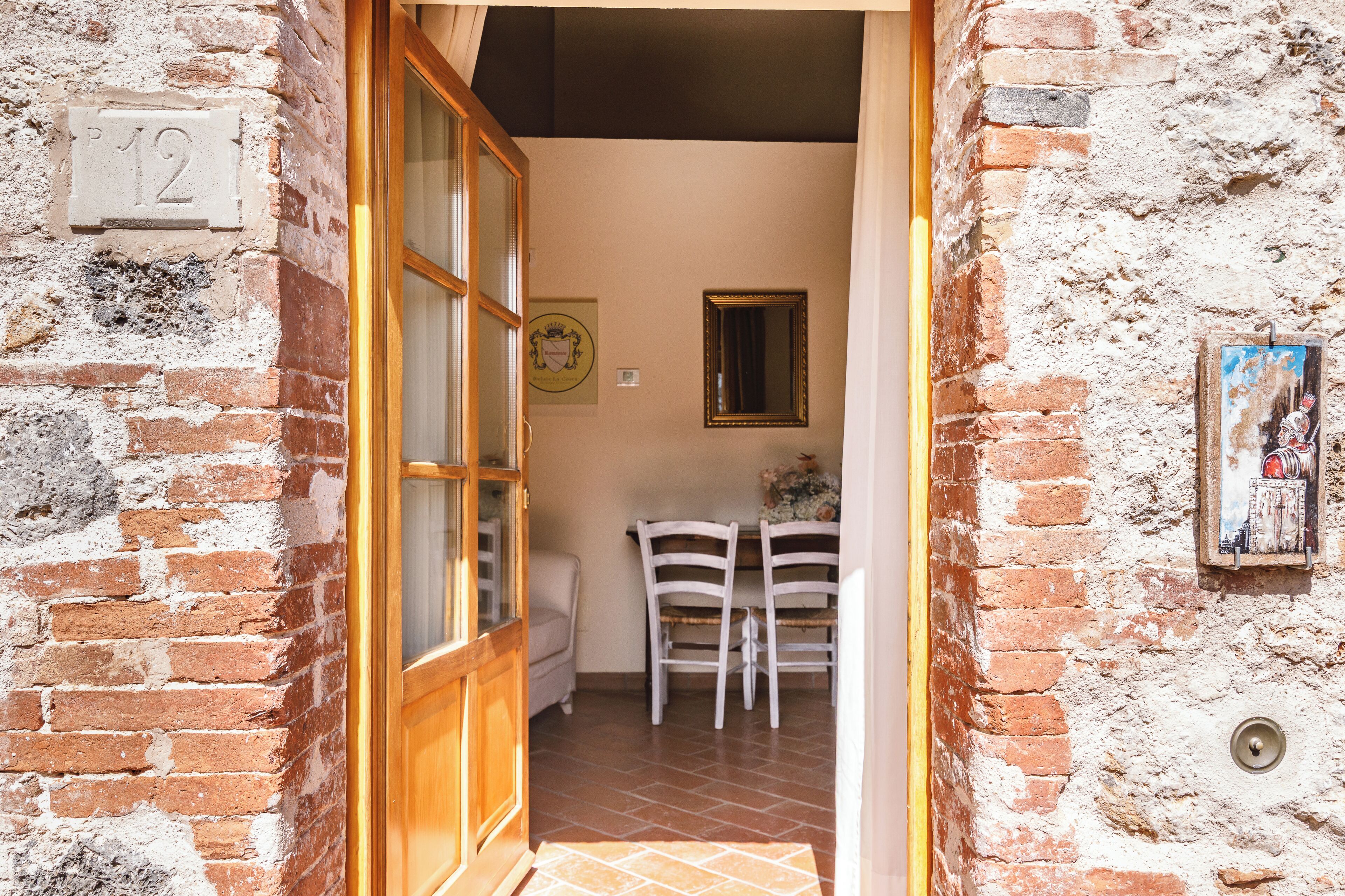 Una habitación rústica con una puerta de madera, una placa con un número, una pequeña mesa con sillas y un cuadro en la pared.