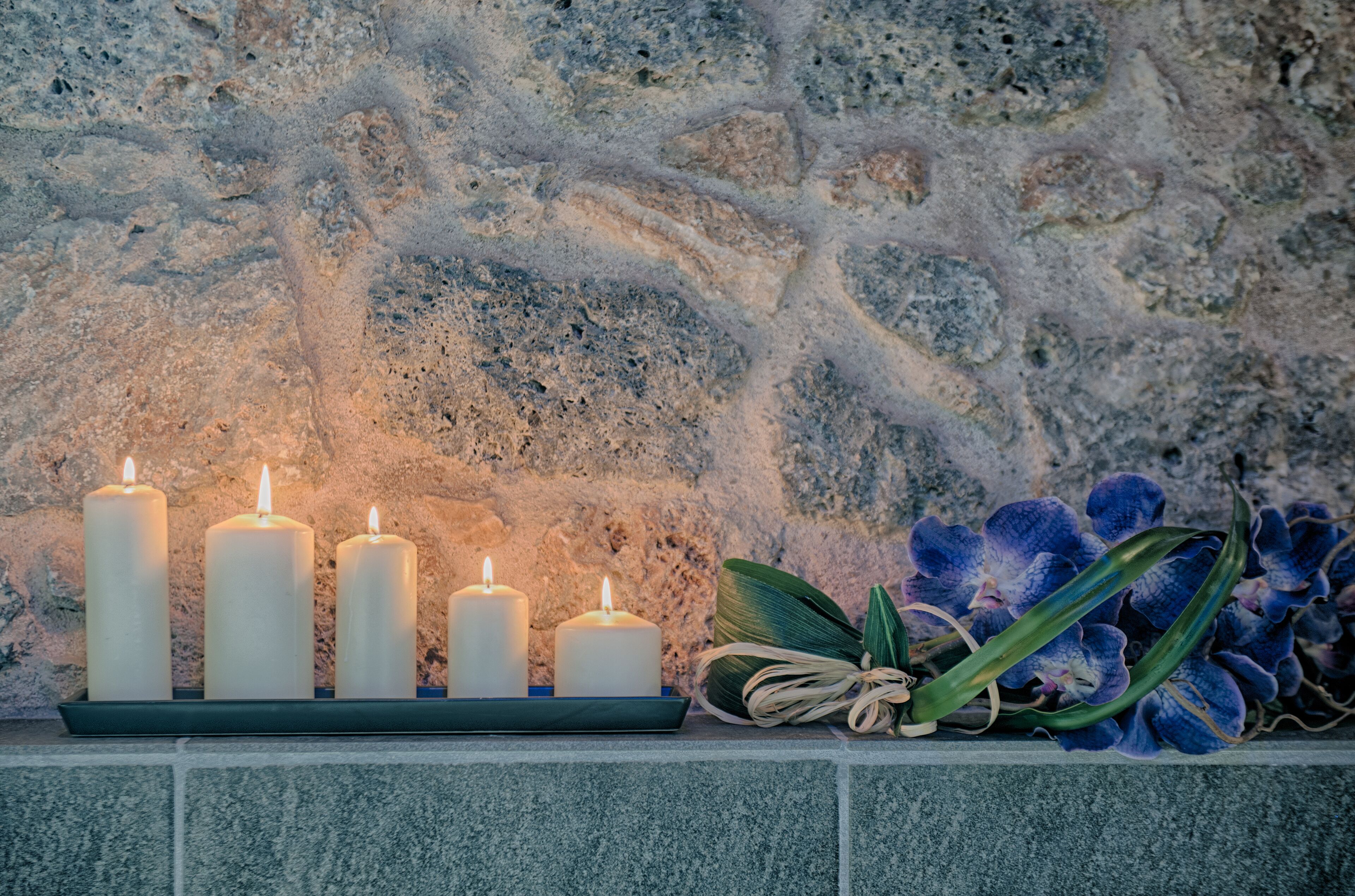 Eine Steinsäule mit fünf brennenden Kerzen auf einer Ablagefläche und einem Strauß blauer Orchideen.