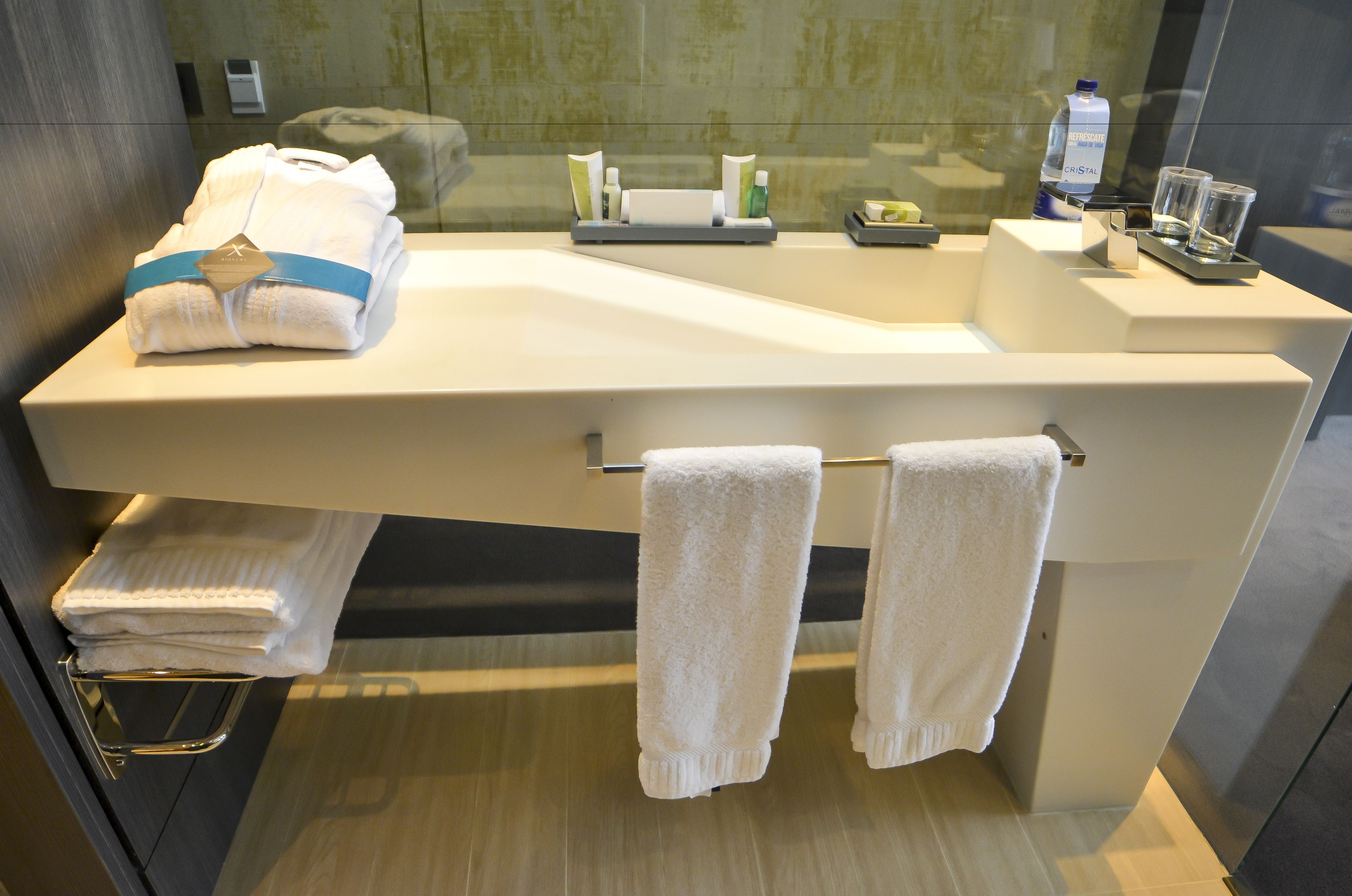 A modern bathroom with a large sink, two towels hanging on a rack, a glass shower enclosure, and toiletries on the counter.