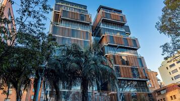 Un immeuble moderne à plusieurs étages, doté de grandes fenêtres et de balcons, entouré de palmiers et d’autres plantes vertes.