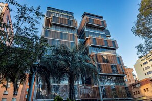 Een modern meerlaags gebouw met grote ramen en balkons, omgeven door palmbomen en andere begroeiing.
