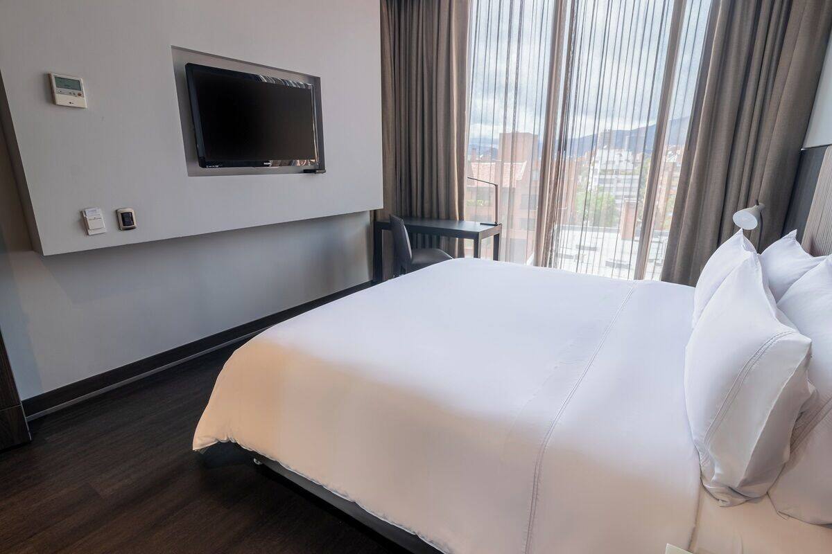 A hotel room with a bed, a TV mounted on the wall, a small table, and a view of the city through large windows.