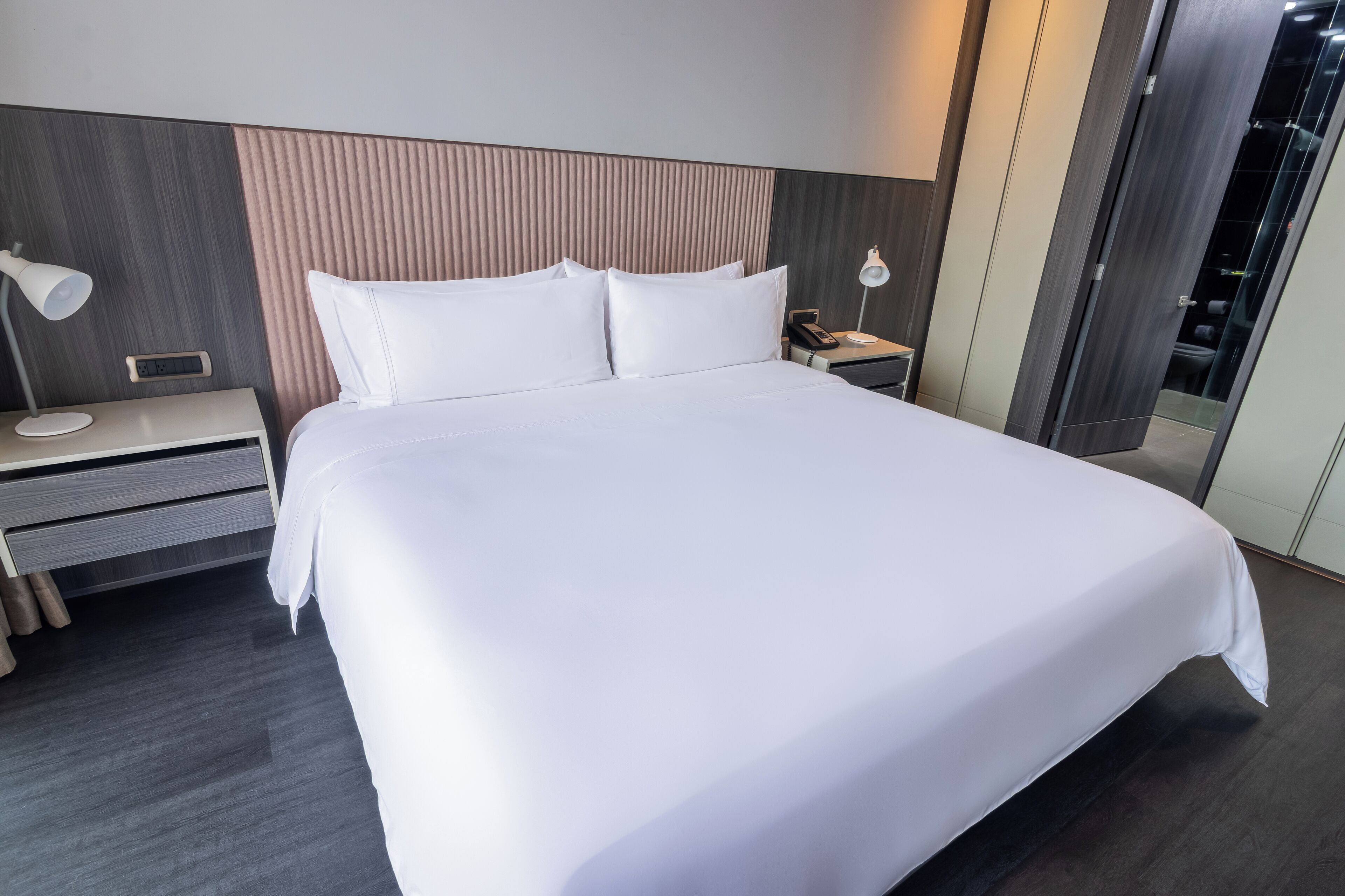 A modern hotel room with a large bed, bedside tables, a nightstand, and a wall-mounted lamp.