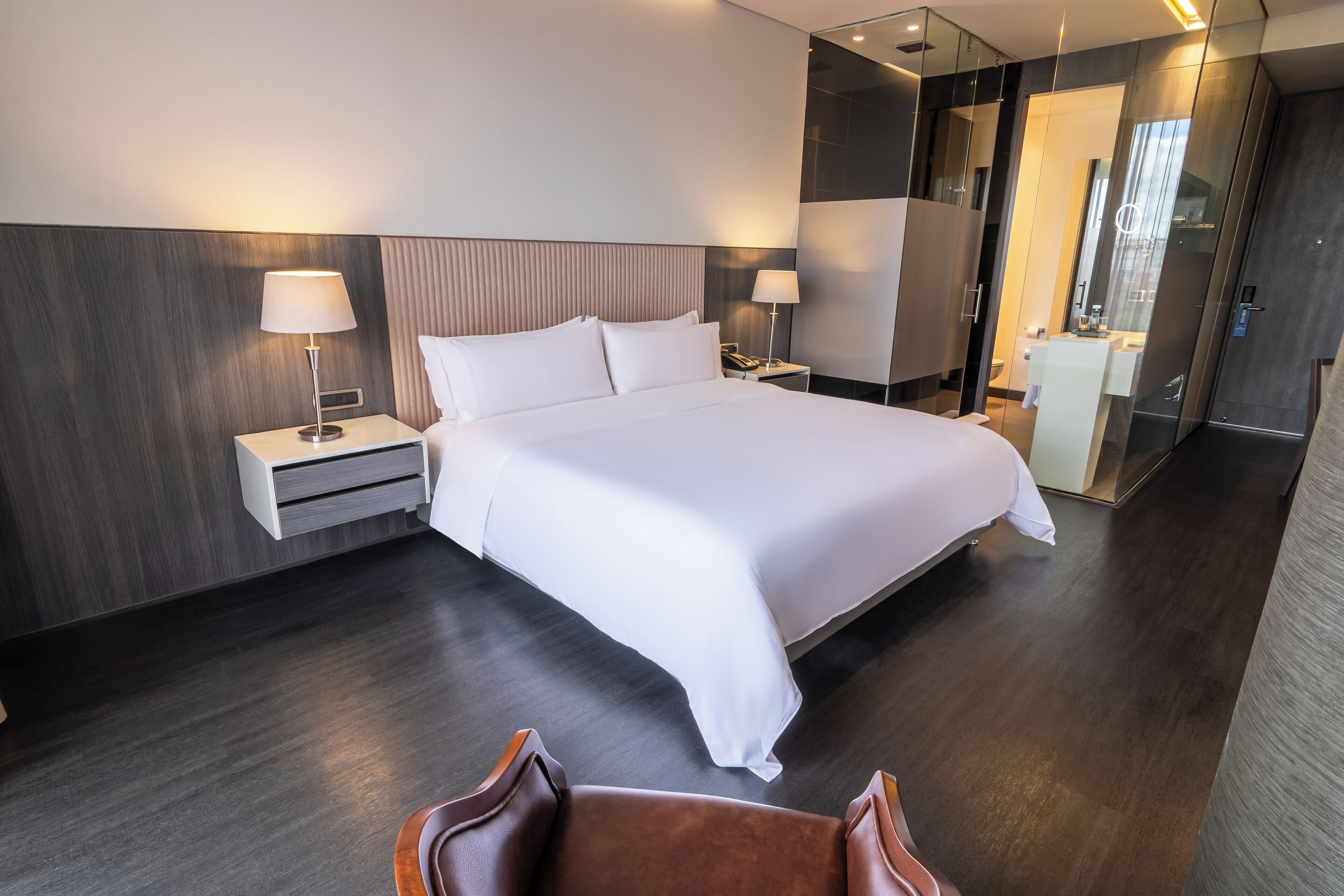 Una habitación de hotel moderna con una cama grande, dos mesitas de noche con lámparas, un cabecero de madera y un box de ducha de vidrio.