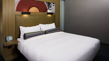 Una cama bien hecha con sábanas blancas y almohadones a rayas, un cabecero de madera y una mesita de noche con un reloj.