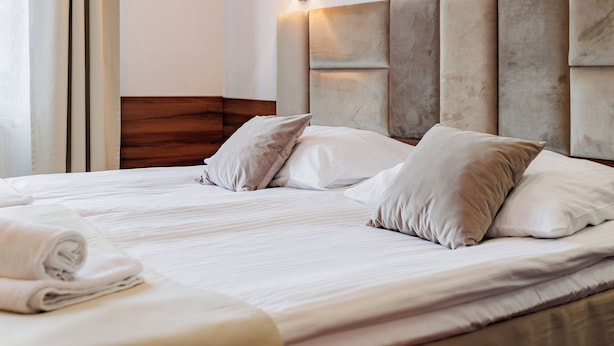 Una cama bien tendida con un cabecero, almohadas y una toalla enrollada.