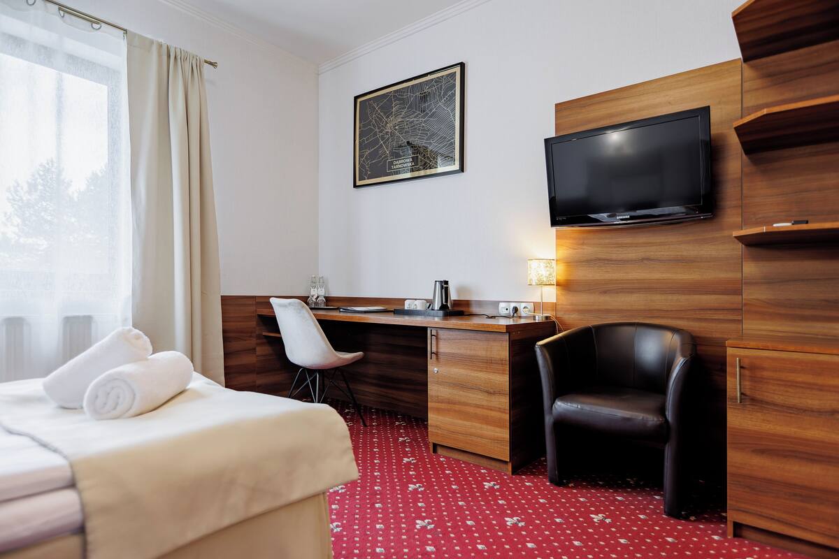 Una habitación de hotel con cama, escritorio, silla, televisor y ventana con cortinas.