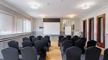 Sala de conferências com fileiras de cadeiras, uma tela de projetor e um grande janelão.