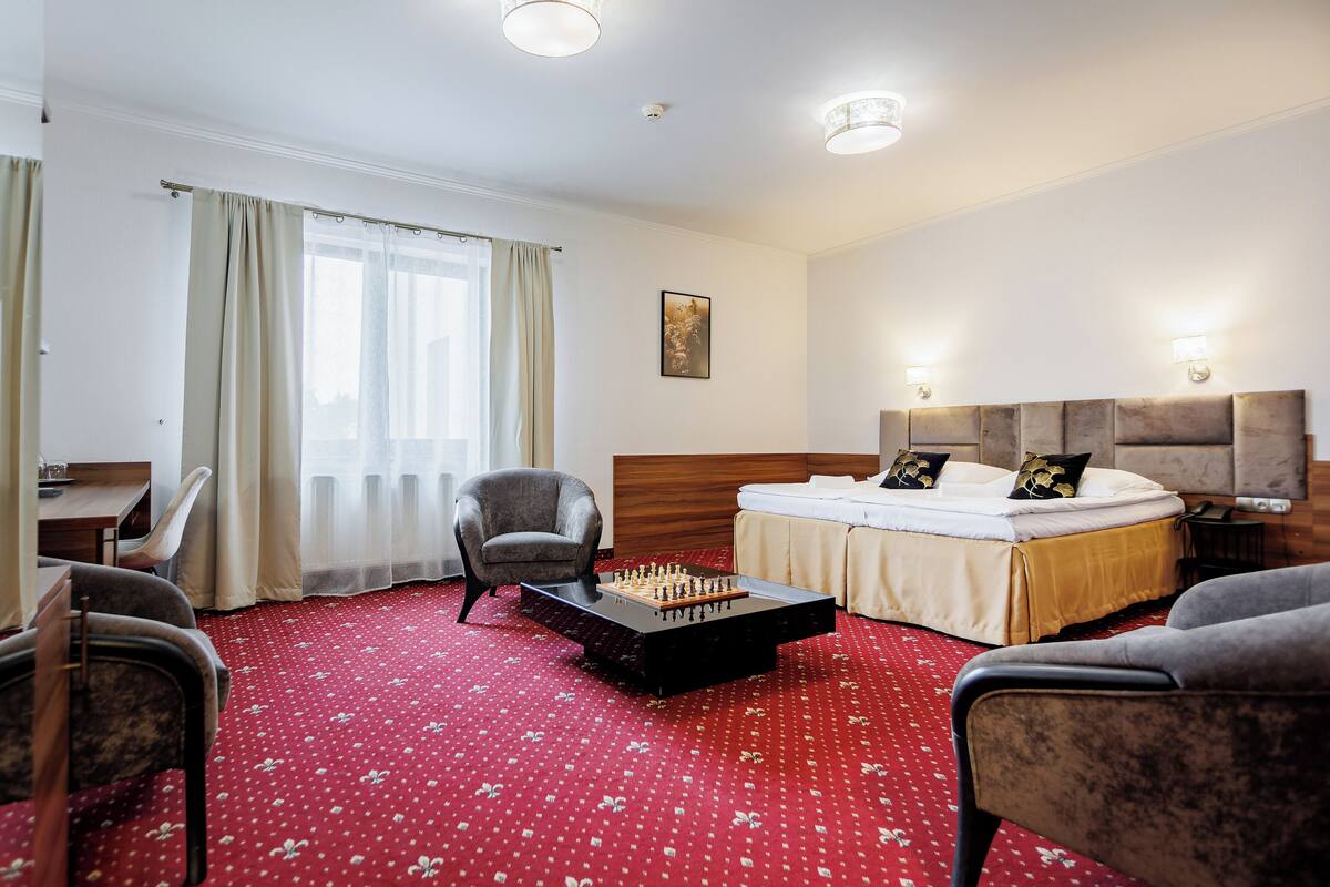 Habitación de hotel con cama, escritorio, silla, mesa auxiliar con un tablero de ajedrez y alfombra roja.
