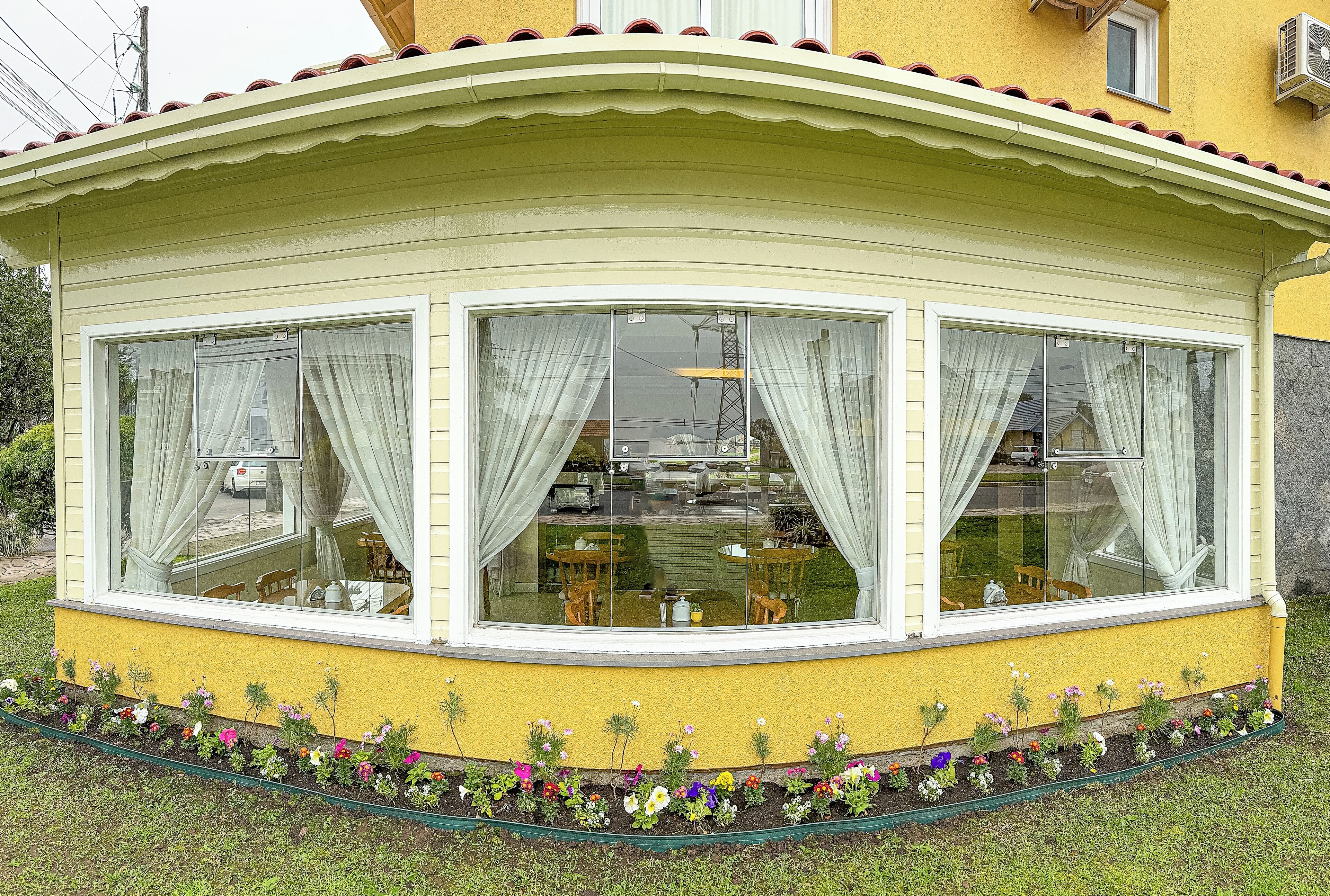 Un edificio giallo con una finestra a bovindo, tende bianche e un'aiuola fiorita di fronte.