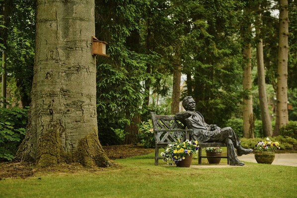 A statue of a seated man on a bench in a park with trees and flower pots. - Cairn Lodge (Auchterarder)