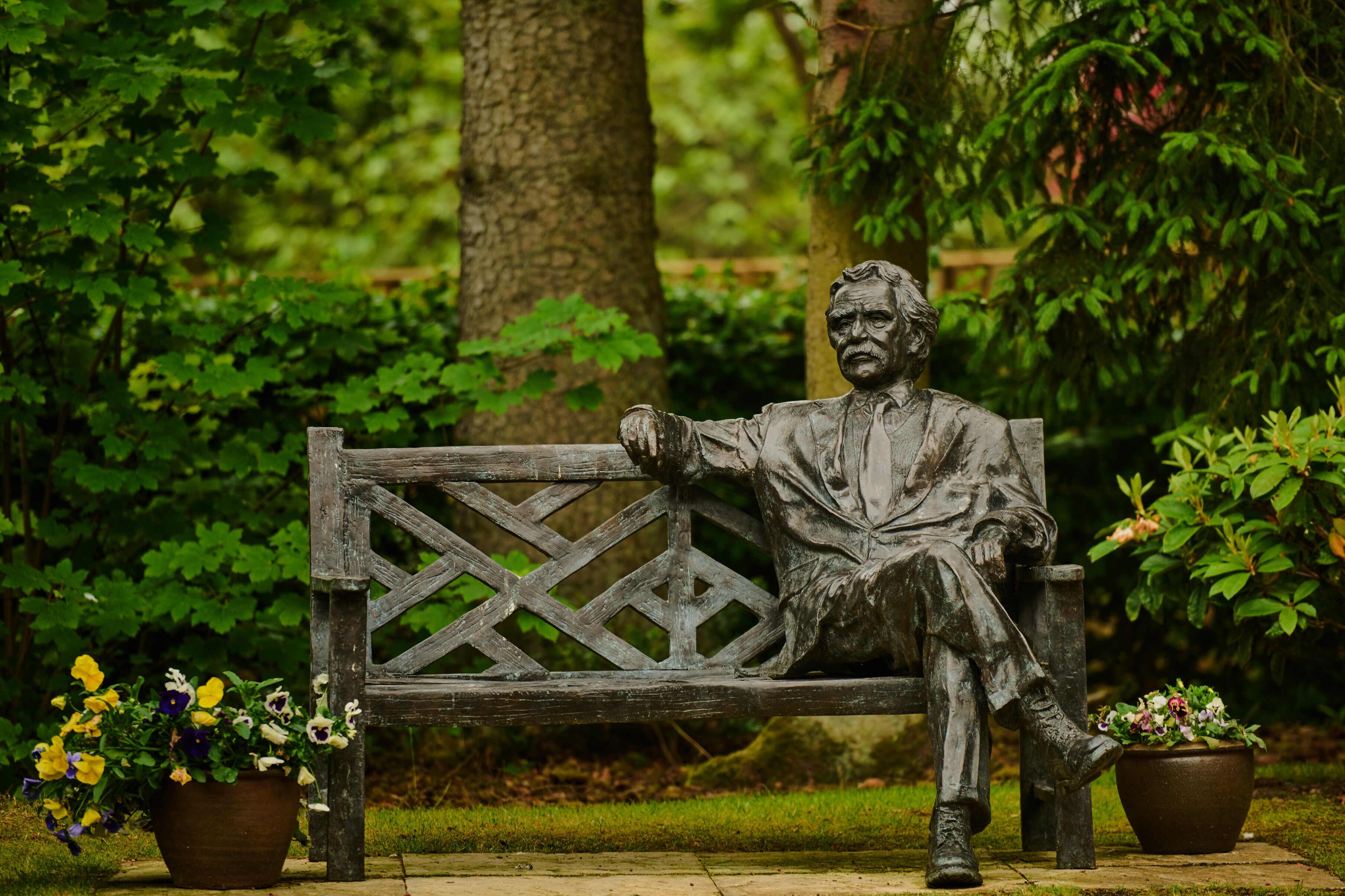 A statue of a seated man on a bench surrounded by greenery.