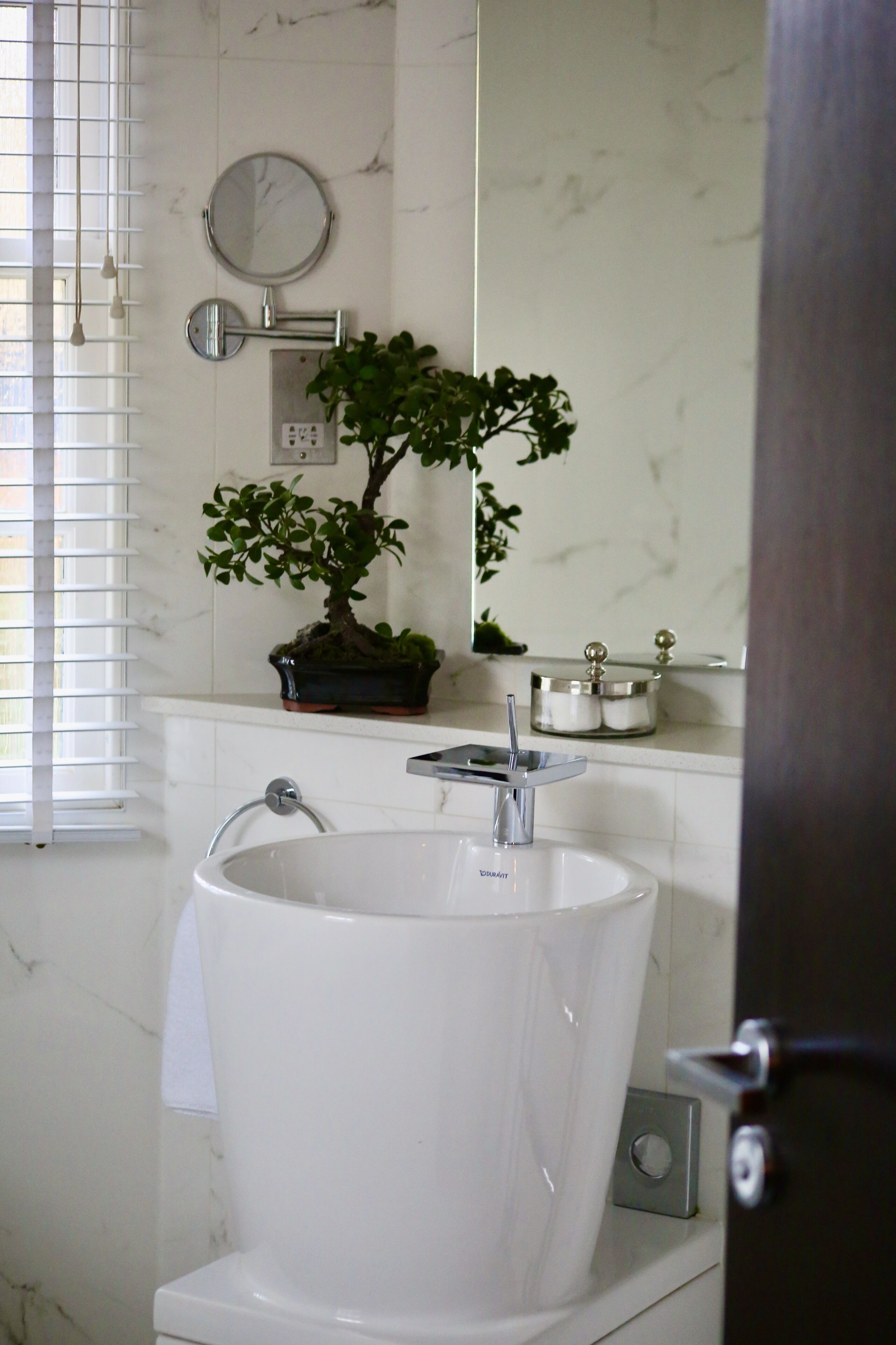 A bathroom with a white sink, a potted plant, a round mirror, and a clock on the wall.