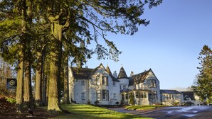 Un grand bâtiment imposant, doté de nombreuses cheminées et d’une entrée remarquable, entouré d’une végétation luxuriante et de grands arbres.