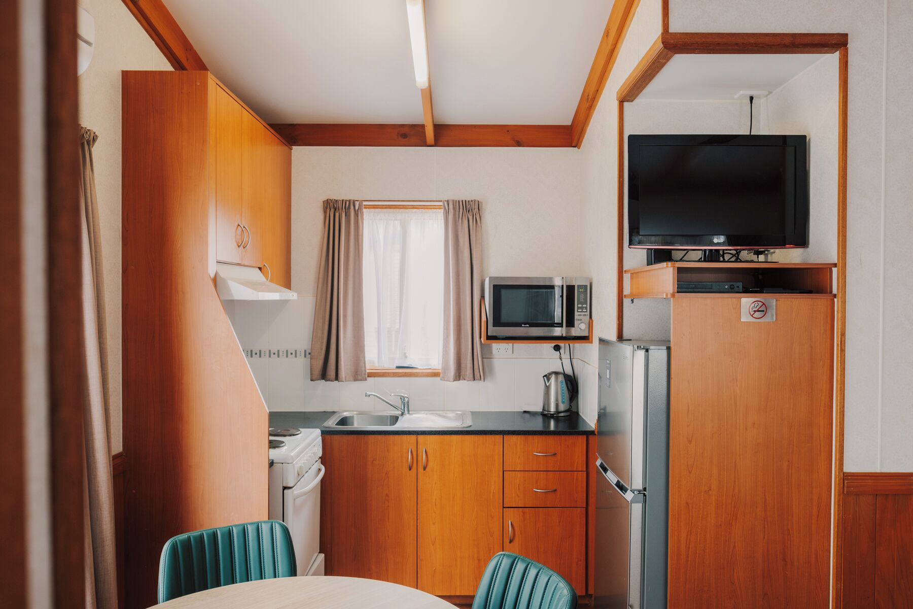 A compact kitchenette with a microwave, stove, sink, and a television on a shelf.