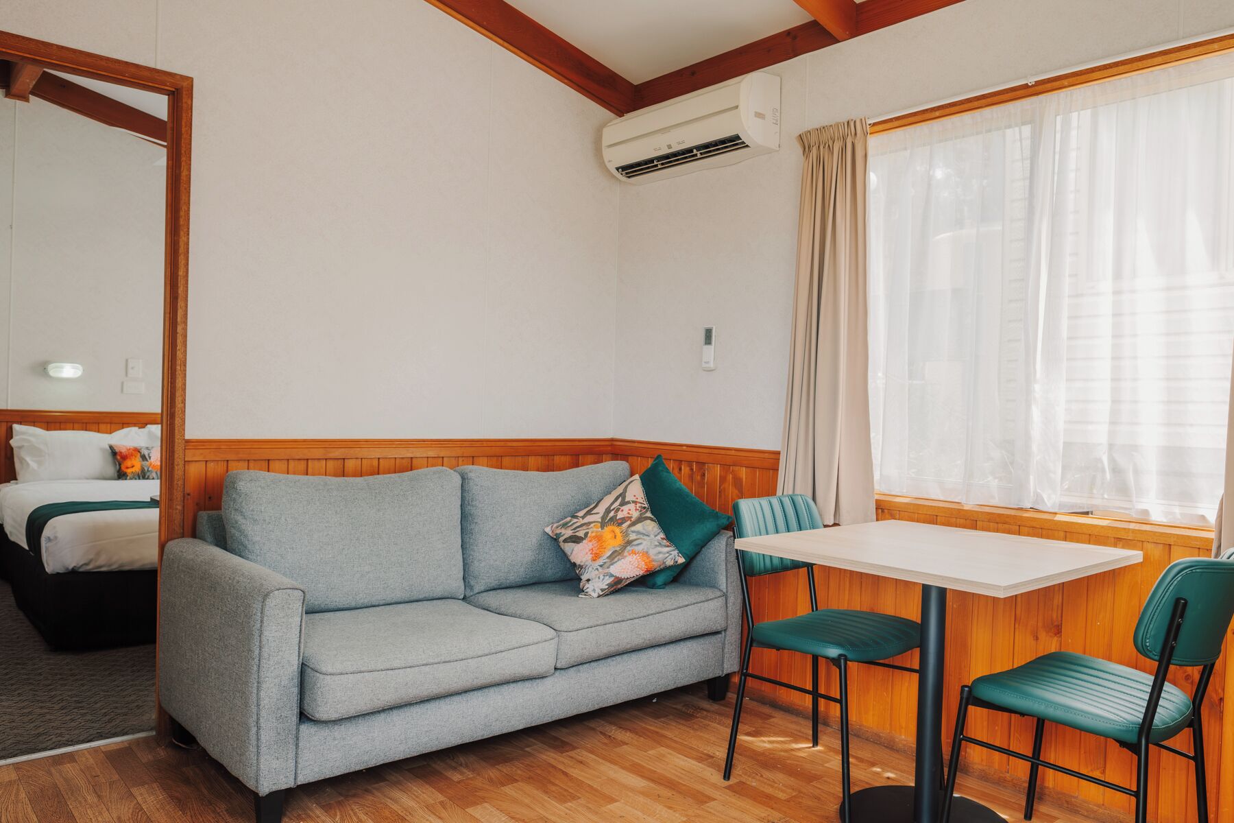 Una habitación con un sofá gris, dos sillas verdes, una mesita, una cama con ropa de cama blanca, un espejo, un equipo de aire acondicionado y una ventana con cortinas translúcidas.