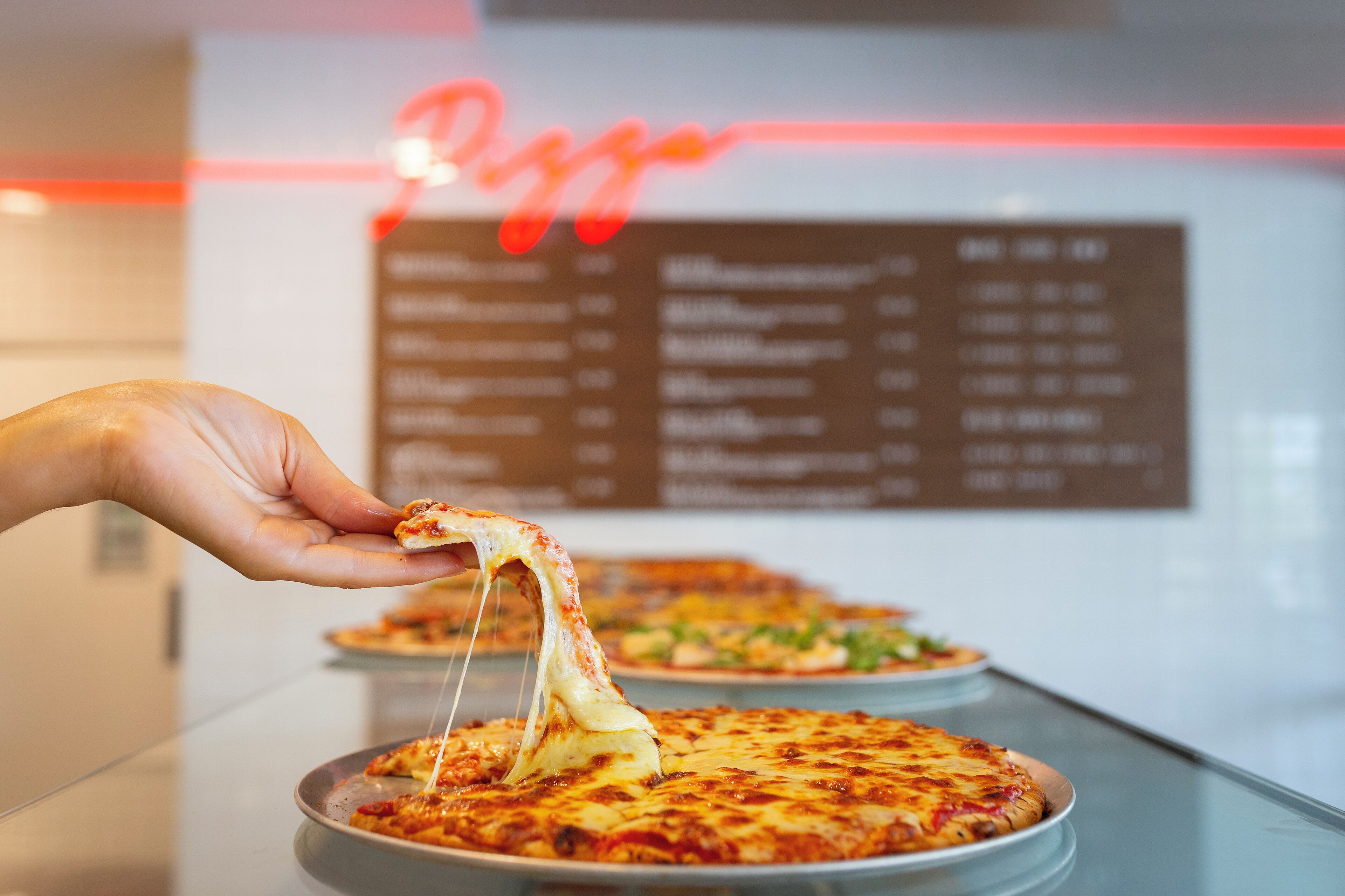 A hand holding a slice of pizza with melted cheese, with a menu board in the background.