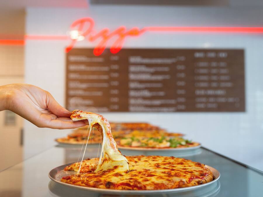 A hand holding a slice of pizza with melted cheese, with a menu board in the background.