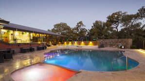 A swimming pool with a curved design, surrounded by a stone wall and a wooden fence. There are outdoor seating areas and a building with large windows and a roof. - Alivio Tourist Park Canberra (O'Connor)
