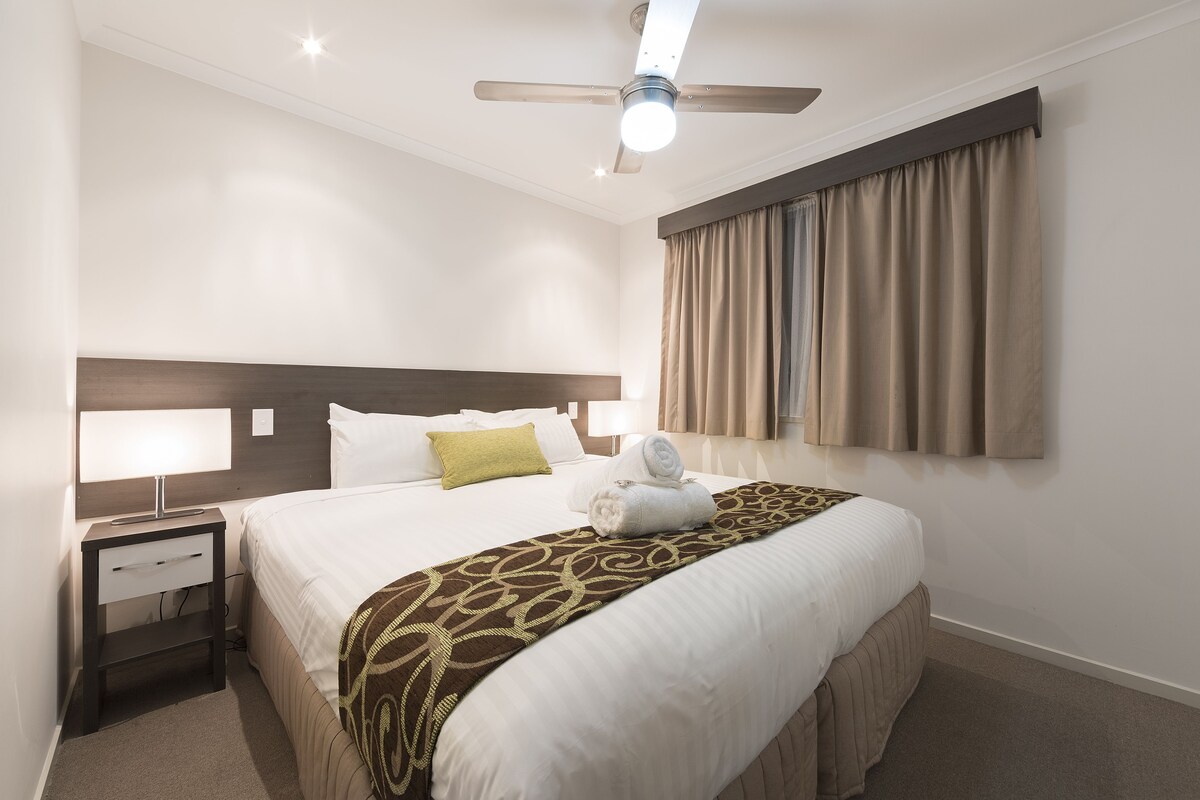 A hotel room with a bed, a nightstand, a ceiling fan, and a window with curtains.