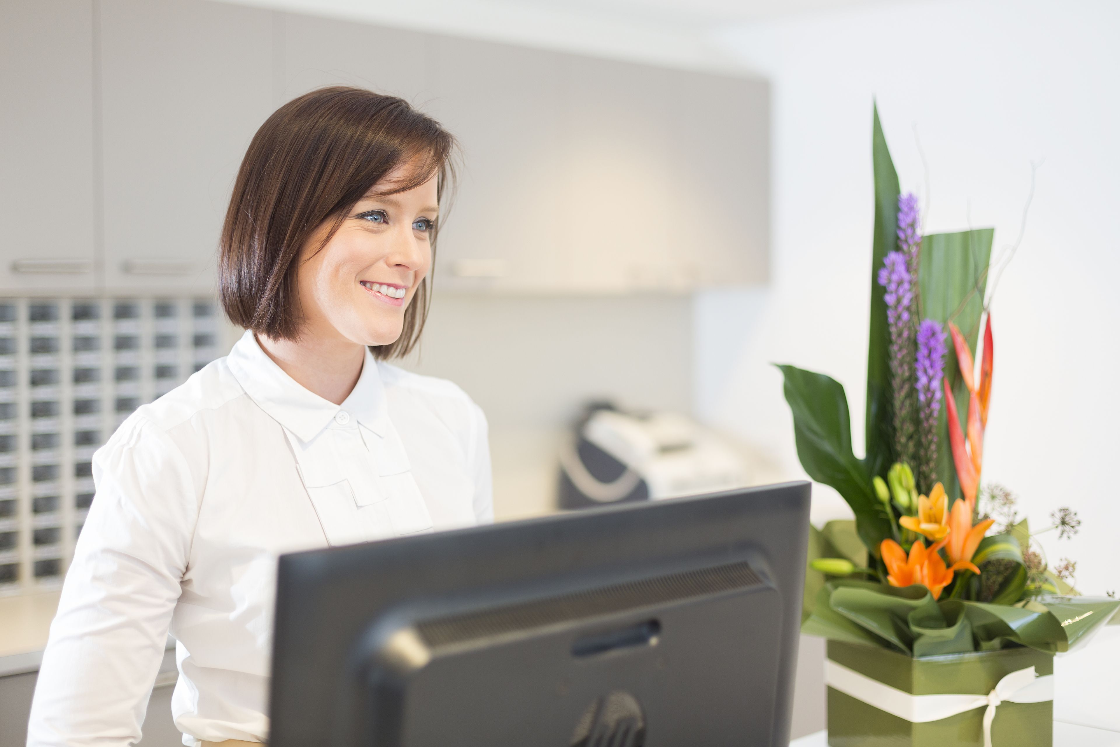 A person sitting at a desk with a computer, a vase of flowers, and a white shirt.