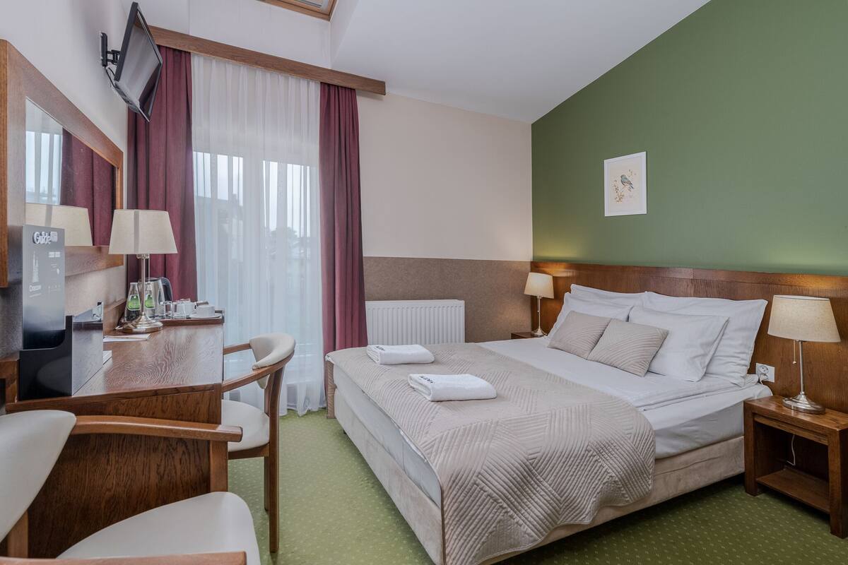 A hotel room with a bed, a desk with a computer, a chair, a lamp, and a window with curtains.