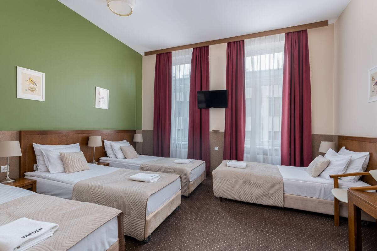 A hotel room with four beds, a television, and large windows with red curtains.