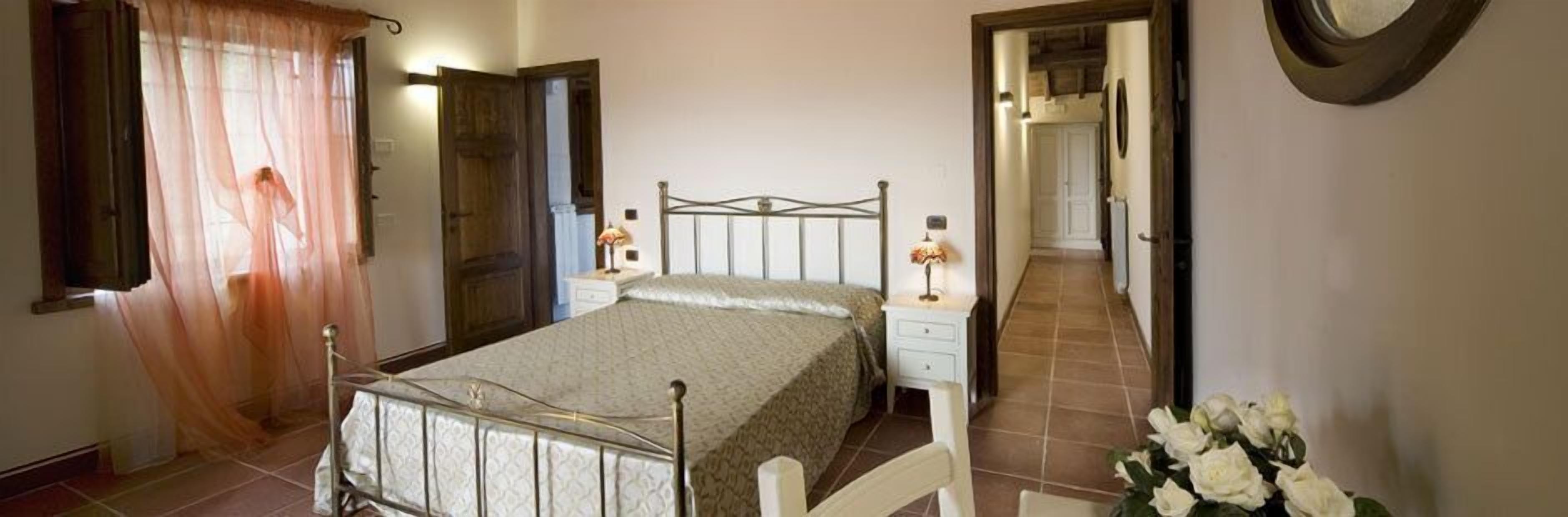 A bedroom with a metal bed frame, a bedside table with a lamp, a window with sheer curtains, and a door leading to another room.