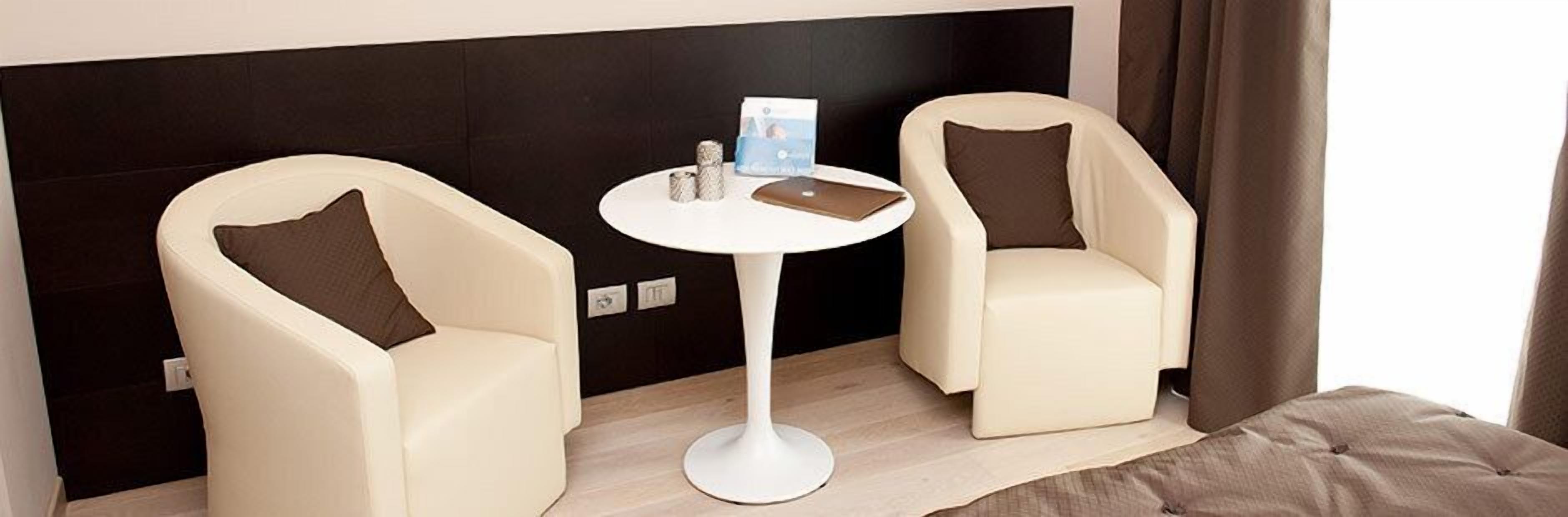 Deux fauteuils blancs avec des coussins marron, une petite table blanche sur laquelle se trouvent un livre et une carte, et un mur sombre avec un interrupteur.