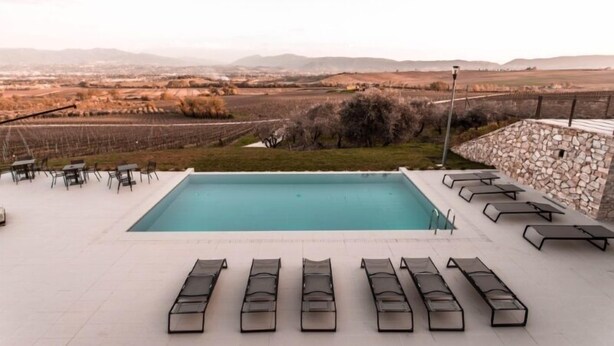 Piscina com espreguiçadeiras e mesas, cercada por um muro de pedra e vista para vinhedos e colinas ao longe.