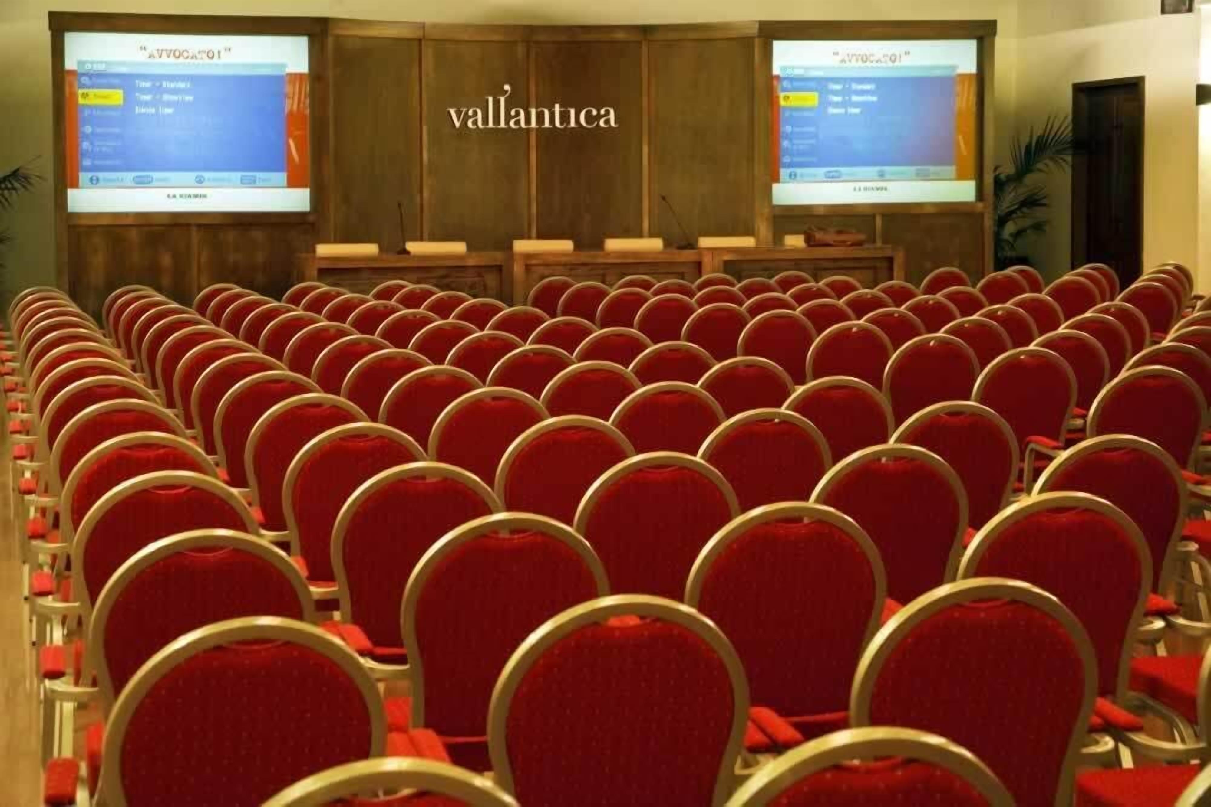 A conference room with rows of red chairs and two projection screens displaying the same content.