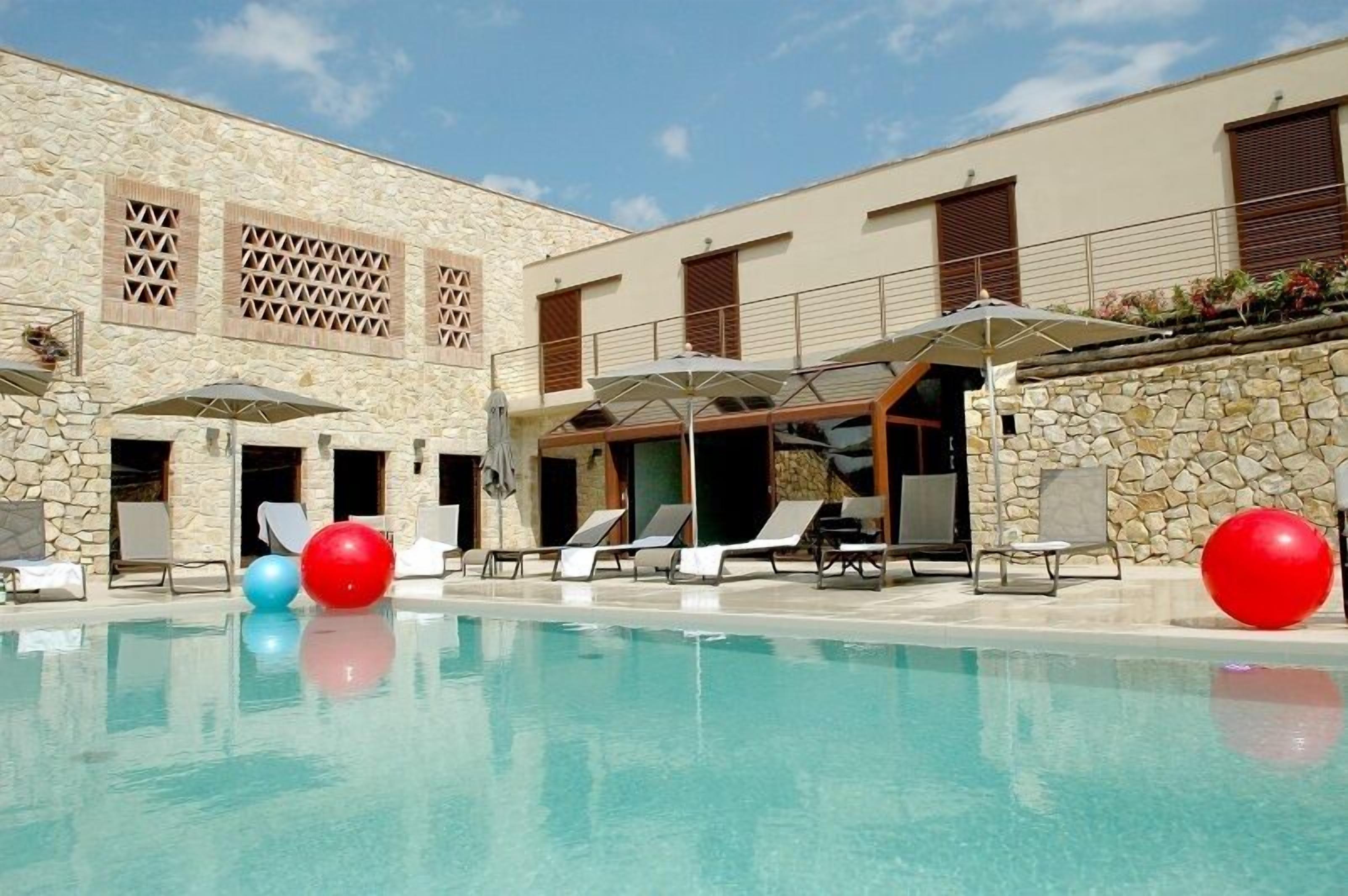 Un espace aménagé au bord de la piscine, avec des chaises longues et des parasols, un bâtiment en pierre doté de volets en bois, et un ciel d’un bleu limpide.
