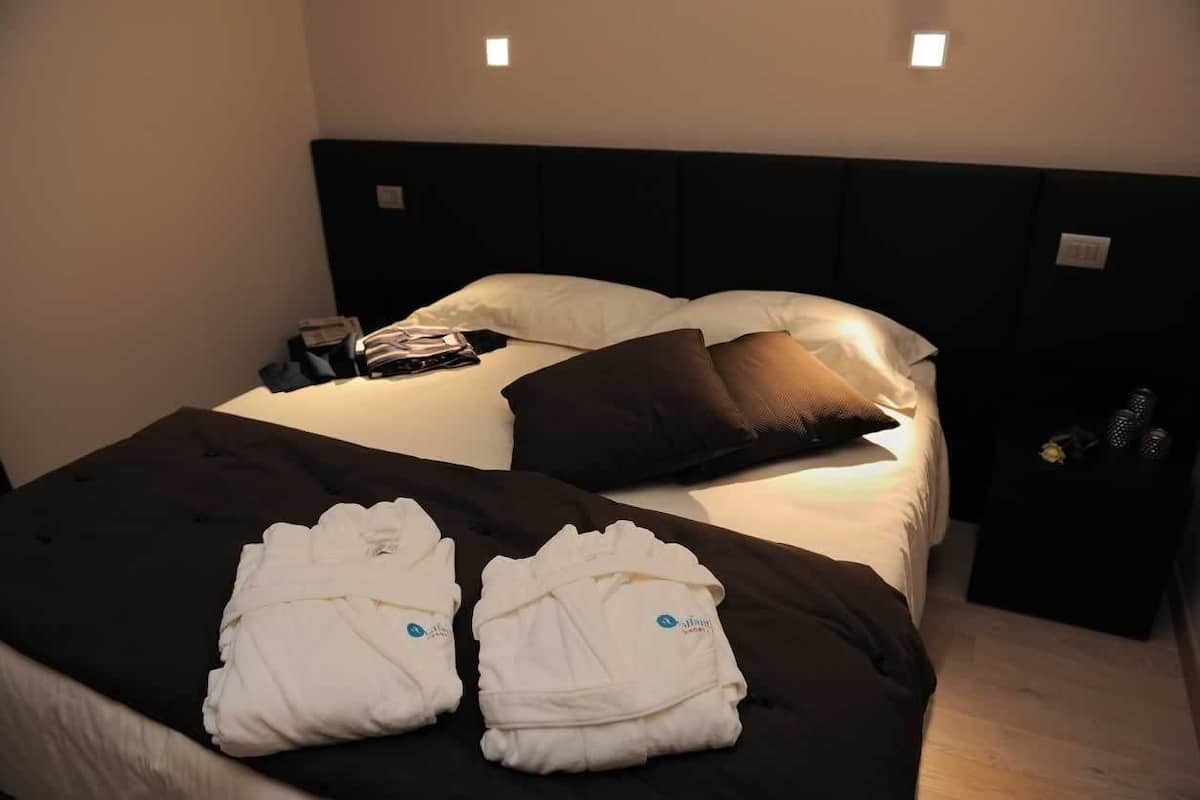 Ein ordentlich bezogenes Bett mit weißer und brauner Bettwäsche, ein kleiner Nachttisch mit Lampe und eine Wandleuchte.