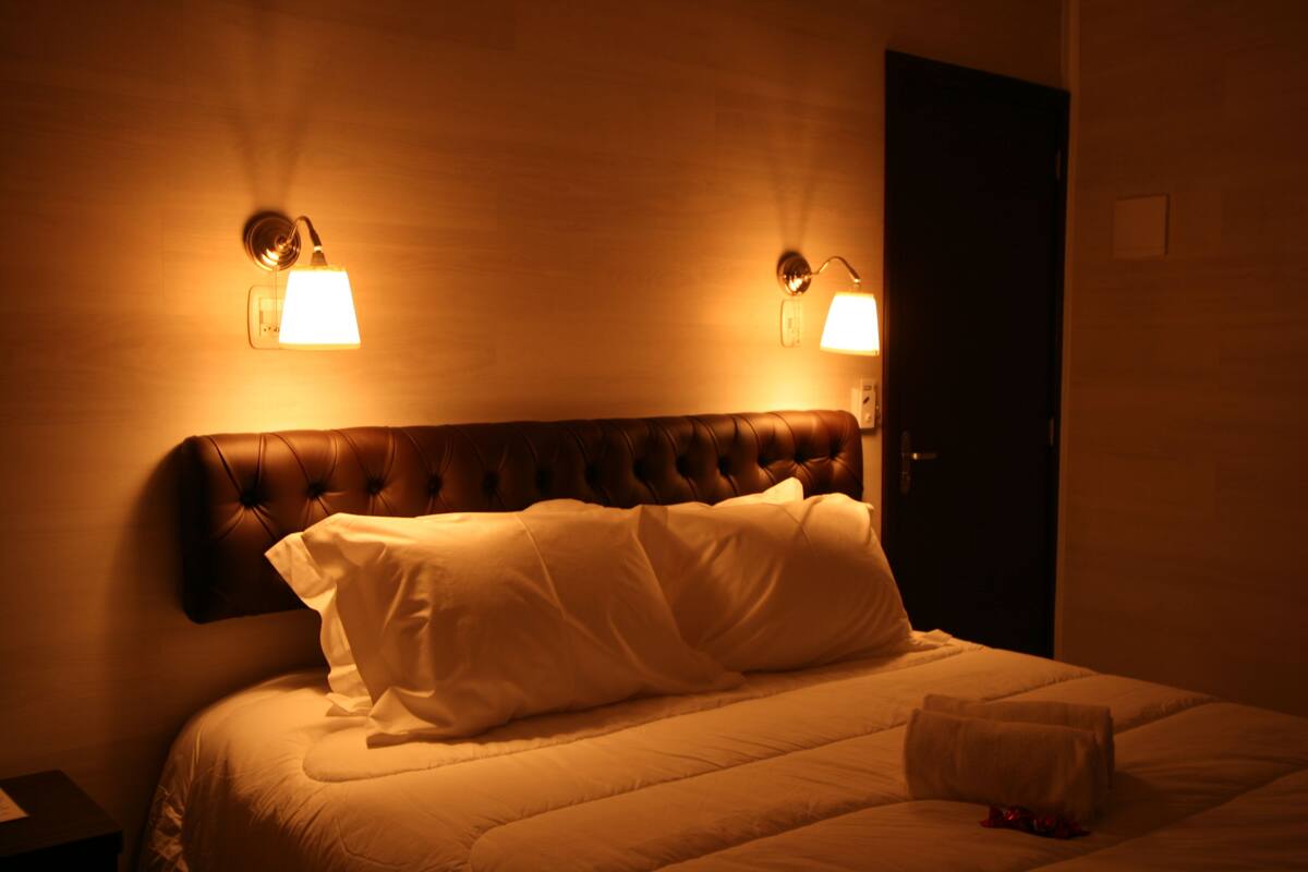 A hotel room with a tufted headboard, two wall-mounted lamps, and a neatly made bed with a folded towel on the side.