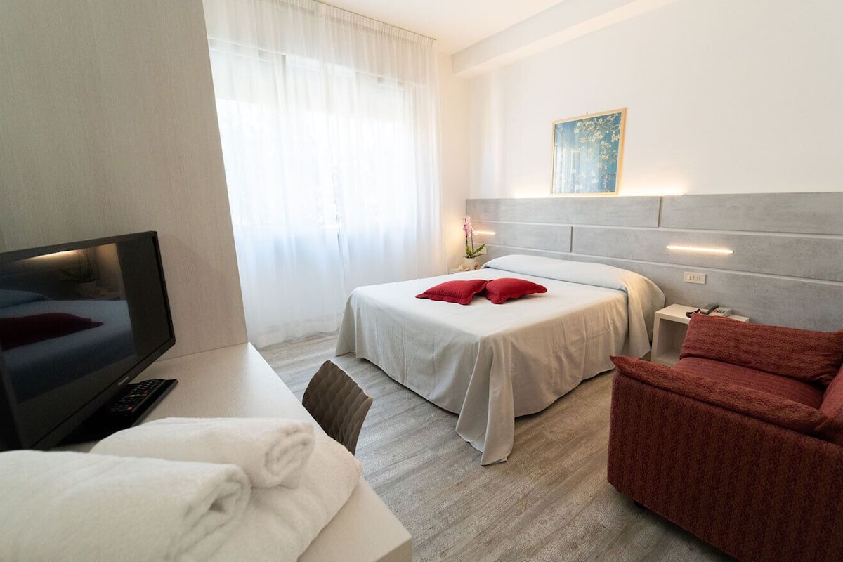 Una habitación de hotel con una cama, un televisor, un sillón rojo y una pequeña mesa con una lámpara.