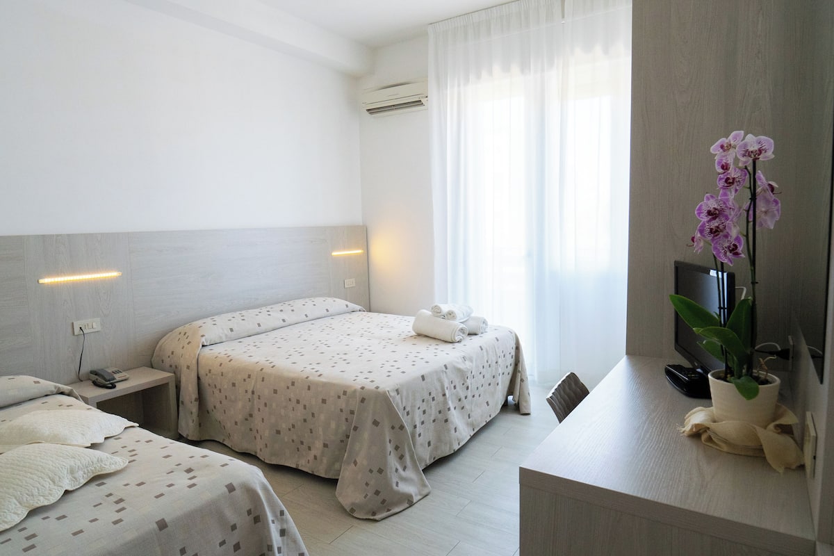 Habitación de hotel con dos camas, una mesita de noche con teléfono, una pequeña mesa con planta y una ventana con cortinas.