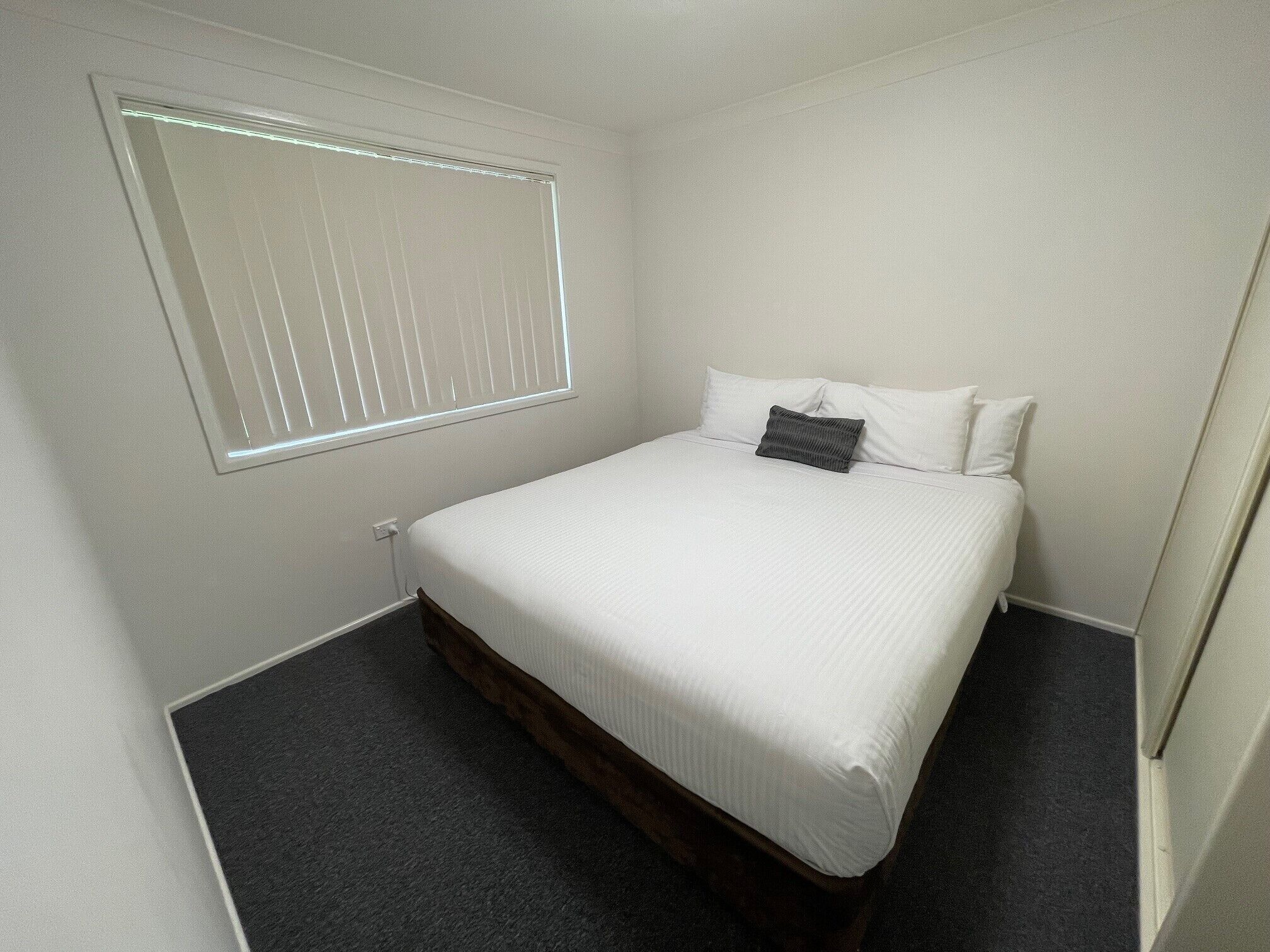 Une petite chambre à coucher, bien rangée, avec un lit simple, une fenêtre avec des stores et un sol recouvert d’une moquette sombre.
