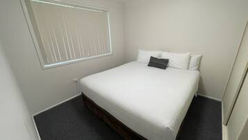 Une petite chambre à coucher, bien rangée, avec un lit simple, une fenêtre avec des stores et un sol recouvert d’une moquette sombre.