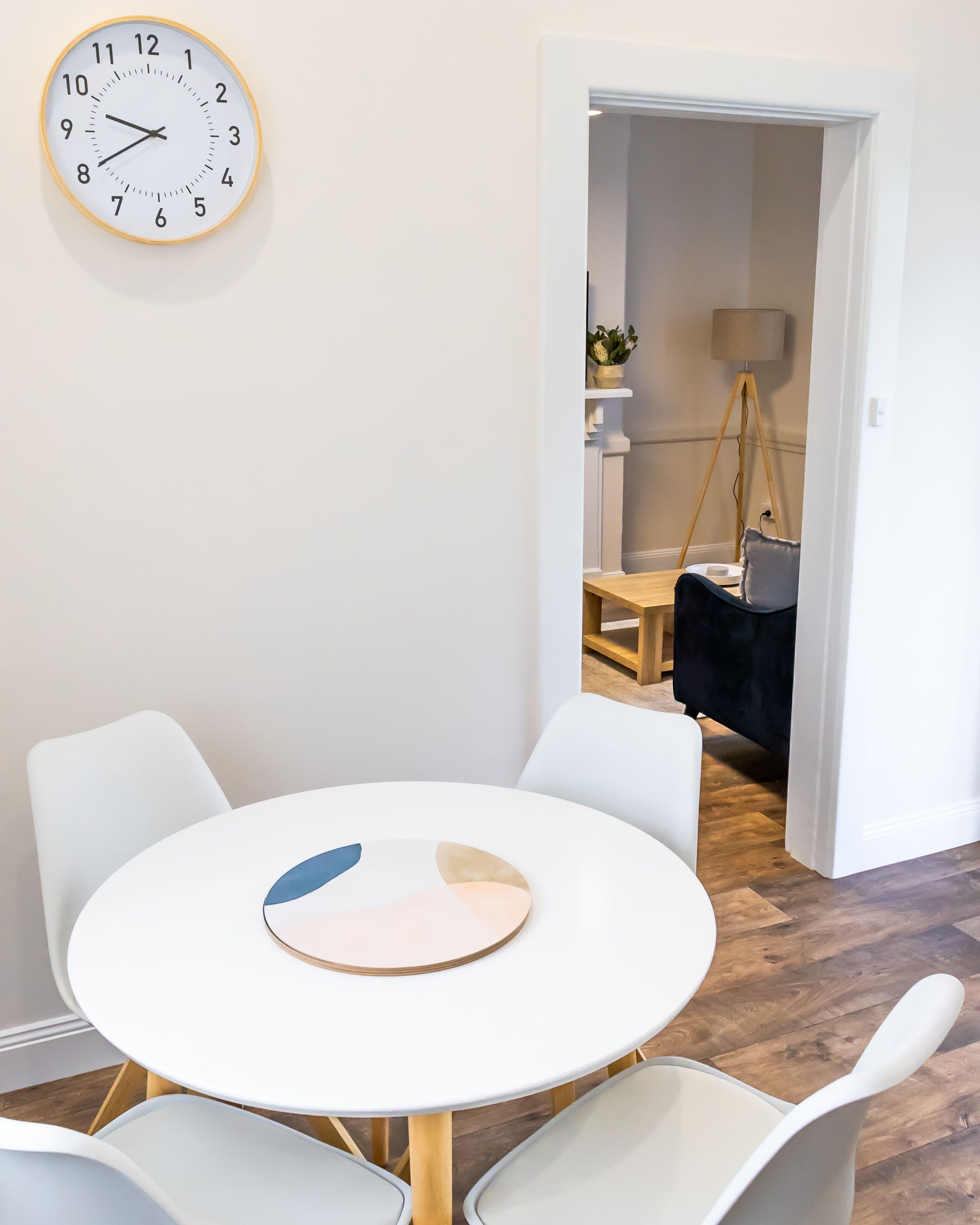 Un comedor moderno con una mesa blanca redonda y cuatro sillas blancas. Un reloj de pared está montado sobre la mesa.